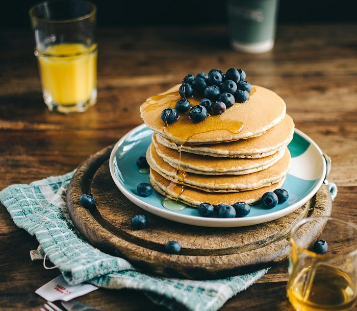 A stack of blueberry pancakes on a wooden table with juice and a jug of syrup