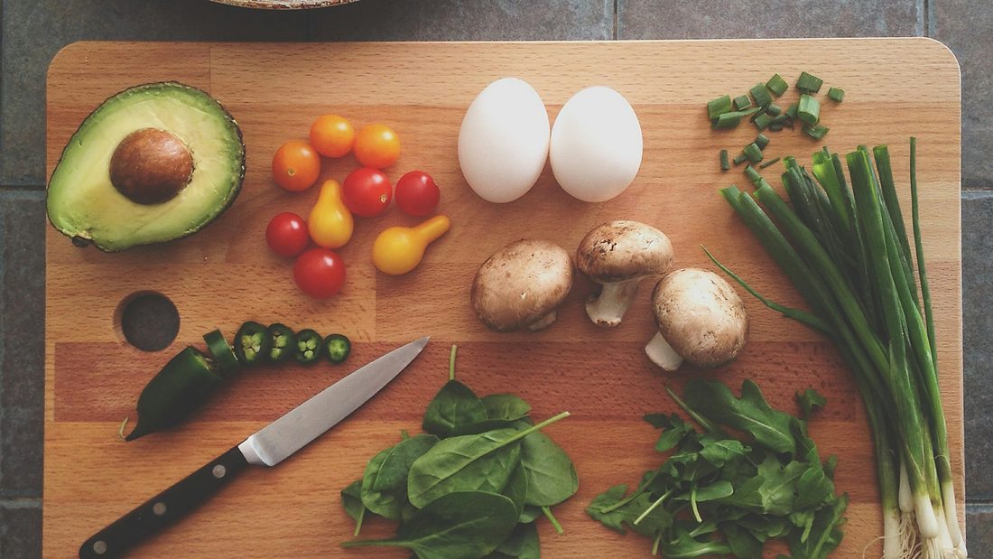 A wooden board with avocado, tomatoes, eggs, mushrooms, spring onions and spinach, beside a knife